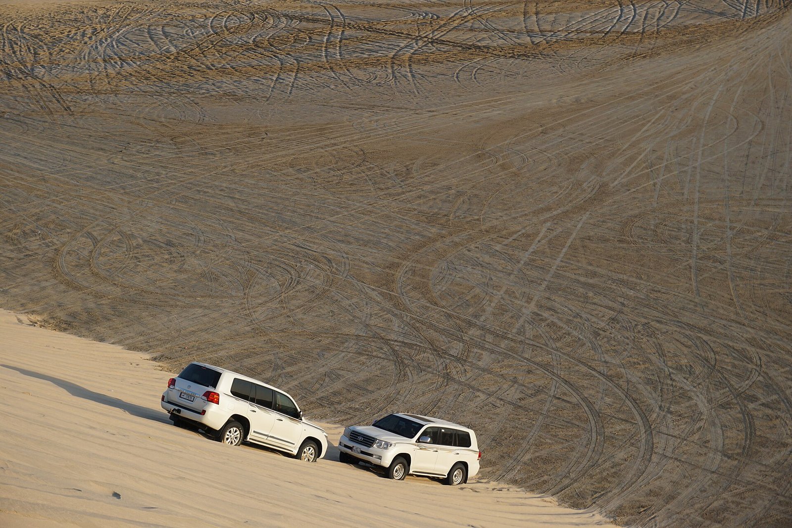 Two 4x4 safari vehicles driving across a steep dune slope in Qatar