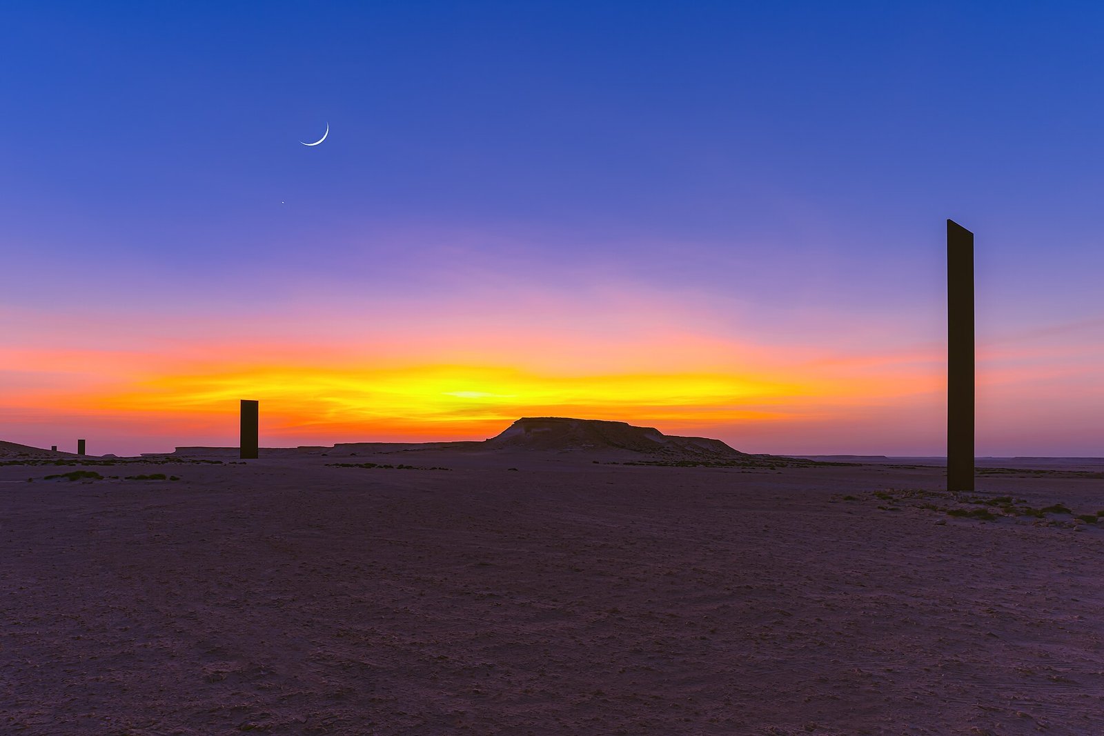 Richard Serra steel plates silhouetted against an orange sunset and crescent moon in the Brouq Nature Reserve